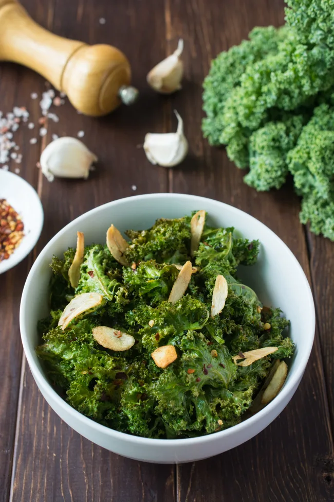 These healthy Spicy Garlic Kale Chips are irresistably crispy and tasty! Fresh kale leaves are tossed in garlic oil and garlic powder, baked till crisp, then topped with crunchy garlic chips and spicy crushed red pepper. 